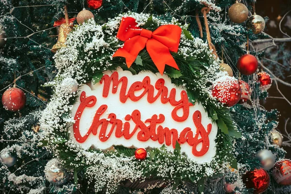 Merry Christmas handwriting logo made from foam on artificial pine tree for christmas time in park. Christmas tree decorations with color lights and merry christmas logo in garden. Selective focus.