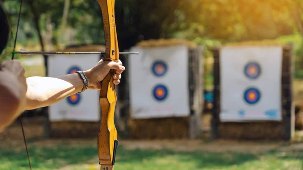 Hands of woman aims archery bow and arrow to colorful target in shooting range during training. Exercise and concentration with outdoor archery. Selective focus on hand. Sport, Recreation concept.