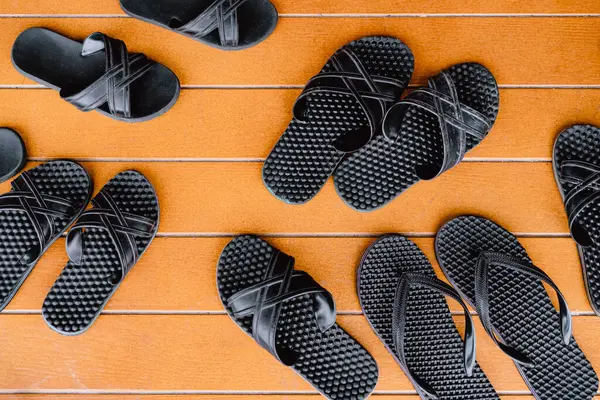 Top view of several pairs of classic black sandals on orange painted wooden background at the patio. Many pairs of rubber flip-flops next to open pool. Hygiene shoes rubber sandals of toilet room.