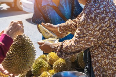 Yaşlı Taylandlı kadın satıcı yol kenarındaki turistlere durian, meyve ve sebze satarak motosiklet sürüyor. Sokak tüccarı taze Durian eti kesip satmış. Durian ticareti. Seçici odak.