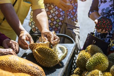 Yaşlı Taylandlı kadın satıcı yol kenarındaki turistlere durian, meyve ve sebze satarak motosiklet sürüyor. Sokak tüccarı taze Durian eti kesip satmış. Durian ticareti. Seçici odak.