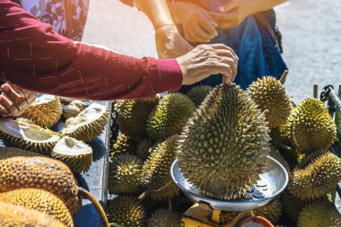 Yaşlı Taylandlı kadın satıcı yol kenarındaki turistlere durian, meyve ve sebze satarak motosiklet sürüyor. Sokak tüccarı taze Durian eti kesip satmış. Durian ticareti. Seçici odak.