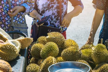 Yaşlı Taylandlı kadın satıcı yol kenarındaki turistlere durian, meyve ve sebze satarak motosiklet sürüyor. Sokak tüccarı taze Durian eti kesip satmış. Durian ticareti. Seçici odak.