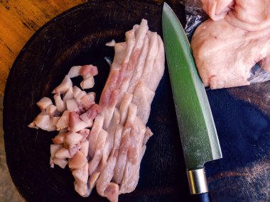 Üst manzara düz, çiğ domuz göbeği ve eski tahta kesme tahtasının üzerinde bıçak. Kesme tahtasında doğranmış çiğ domuz eti ve siyah arka planda bıçak. Esmer arka planda yemek pişirmek için et bıçağı.