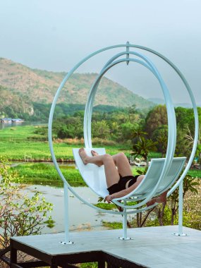 Nehir kıyısındaki tepede doğa manzaralı rahat koltukta dinlenen mutlu kadın. Dişi, yamaçtaki terasta kıvrımlı koltuklarda dinleniyor. Arka planda dağlar ve nehir var..