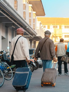 Günlük giysiler içinde bavul taşıyan yaşlı bir gezginin arka görüntüsü. Kıdemli adam sabah bavuluyla sokakta yürüyor. Tatil için bagajı olan bir erkek turist. Tatil ve seyahat kavramı.