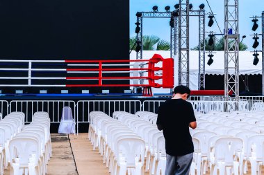Young man using smart phone in front of outdoors boxing ring and many chairs for spectators prepared for competition. Male officer with  empty boxing ring for competition. Boxing ring with white seats clipart