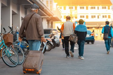 Günlük giysiler içinde bavul taşıyan yaşlı bir gezginin arka görüntüsü. Kıdemli adam sabah bavuluyla sokakta yürüyor. Tatil için bagajı olan bir erkek turist. Tatil ve seyahat kavramı.