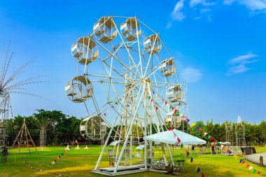 Yeşil çimenlikteki festival ve karnaval standında eğlenmek için beyaz dönme dolaba ışık ve küçük bayraklarla süslenmiş güzel bir manzara. Arka planda güzel doğa olan açık hava tema parkı