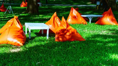 Beautiful view of orange triangle bean bags and cushions with white wooden table for customers to relax outdoors on green grass under shade of large tree with blurred background. Selective focus.