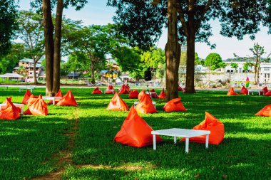 Beautiful view of orange triangle bean bags and cushions with white wooden table for customers to relax outdoors on green grass under shade of large tree with blurred background. Selective focus.