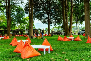 KANCHANABURI, THAILAND - 13 Temmuz 2020: had Chuen Ruedee 'deki ağacın gölgesinde yeşil çimlerin üzerinde dinlenmeleri için beyaz ahşap masalı turuncu üçgen fasulye torbaları ve yastıkların görüntüsü.