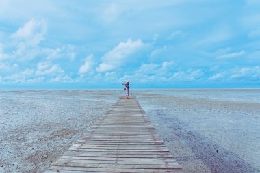 PHETCHABURI-THAILAND, 25 Temmuz 2020: Tanımlanamayan mutlu Asyalı kadın tropikal sahil iskelesinde dinleniyor (ahşap iskele) ve arka planda alçak okyanus gelgiti ile Kral 'ın Kraliyet Girişimi Laem Phak Bia.