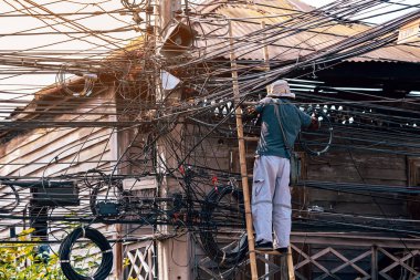 Elektrikçi, eski bir ahşap köyde elektrik sistemini onarmak için merdivenin üzerinde duruyor. Elektrik direklerine bağlı fiber optik kablolar için merdivende çalışan teknisyen..