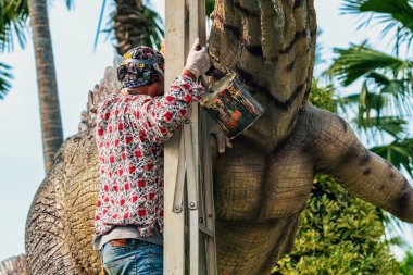 PATTAYA, THAILAND-Aralık 30. 2018: Ressam, Nong Nooch Tropik Botanik Bahçesi 'ni güzel ve gerçekçi kılmak için büyük dinozor heykeli veya heykelini boyamak ve dekore etmek üzere merdivenin üzerinde duruyor.
