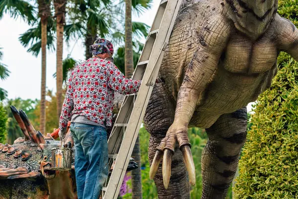 PATTAYA, THAILAND-Aralık 30. 2018: Ressam, Nong Nooch Tropik Botanik Bahçesi 'ni güzel ve gerçekçi kılmak için büyük dinozor heykeli veya heykelini boyamak ve dekore etmek üzere merdivenin üzerinde duruyor.