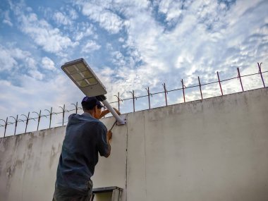 Asyalı yaşlı elektrikçi çimento ya da beton duvar arka planına güneş enerjili hareket sensör ışığı yerleştiriyor. Yaşlı adam, güneş ışığını güzel gökyüzü olan duvarlara kur ya da monte et.