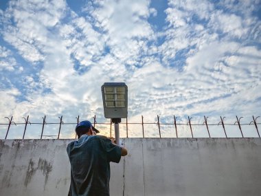 Asyalı yaşlı elektrikçi çimento ya da beton duvar arka planına güneş enerjili hareket sensör ışığı yerleştiriyor. Yaşlı adam, güneş ışığını güzel gökyüzü olan duvarlara kur ya da monte et.