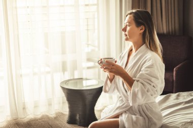 Woman in white bathrobe lying on sofa and relaxing with orange cocktail at the roof of hotel