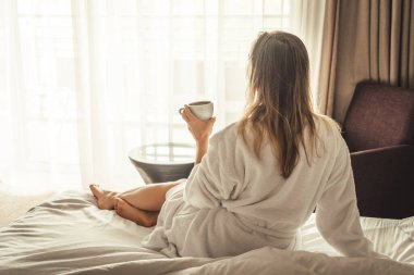 Woman in white bathrobe lying on sofa and relaxing with cup of tea at home near window