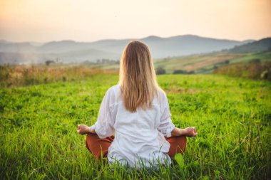 Dağın tepesindeki yeşil çimlerde yoga yapan kadın gün batımında güzel manzaralı.