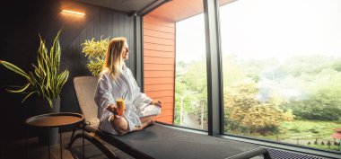 Woman in white bathrobe lying on sofa and relaxing with orange cocktail at home near window