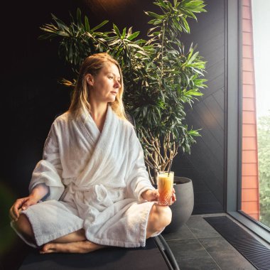 Woman in white bathrobe lying on sofa and relaxing with orange cocktail at home near window