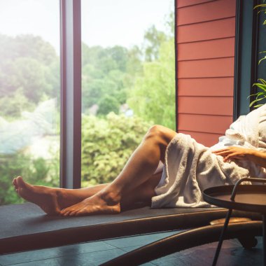 Woman in white bathrobe lying on sofa and relaxing with at home near window