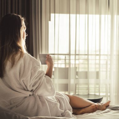 Woman in white bathrobe lying on sofa and relaxing with cup of tea at home near window