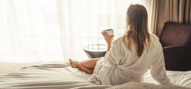 Woman in white bathrobe lying on sofa and relaxing with cup of tea at home near window
