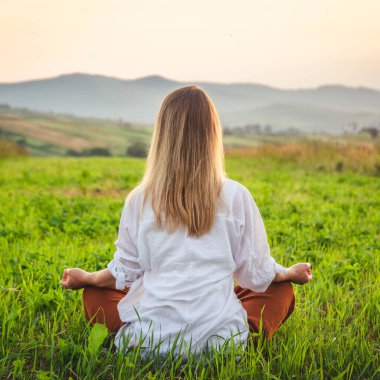 Dağın tepesindeki yeşil çimlerde yoga yapan kadın gün batımında güzel manzaralı.