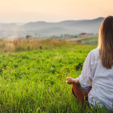 Dağın tepesindeki yeşil çimlerde yoga yapan kadın gün batımında güzel manzaralı.