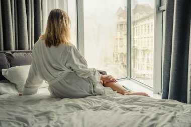 Woman in white bathrobe lying on sofa and relaxing with at home near window