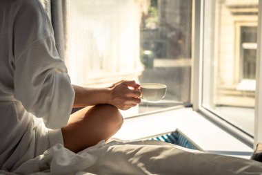 Woman in white bathrobe lying on sofa and relaxing with cup of tea at home near window