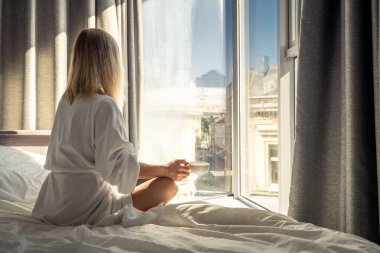 Woman in white bathrobe lying on sofa and relaxing with cup of tea at home near window