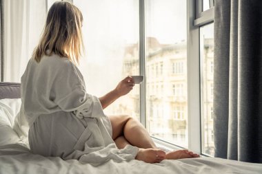 Woman in white bathrobe lying on sofa and relaxing with cup of tea at home near window