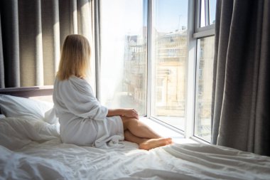 Bornozlu genç bir kadın yatakta oturup otelin penceresinden dışarı bakıyor.