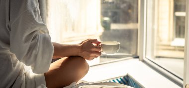 Woman in white bathrobe lying on sofa and relaxing with cup of tea at home near window