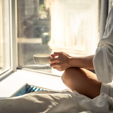 Woman in white bathrobe lying on sofa and relaxing with cup of tea at home near window