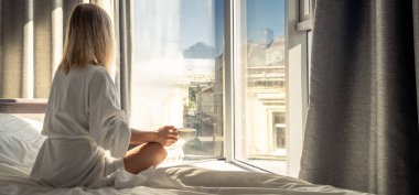 Woman in white bathrobe lying on sofa and relaxing with cup of tea at home near window