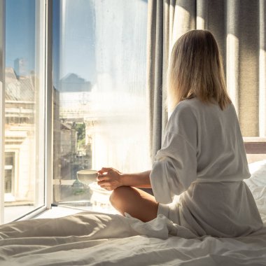 Woman in white bathrobe lying on sofa and relaxing with cup of tea at home near window