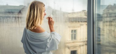 Zarif bornozlu kadın otel odasında kahve içiyor ve pencerenin yanında duruyor. Pencere yansıması