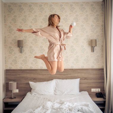 beautiful female droping pilows and jumping with bathrobe on bed in hotel room