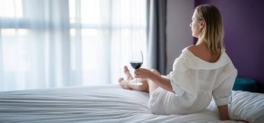 Beyaz bornozlu kadın kanepede uzanıyor ve pencerenin yanındaki otelde bir bardak şarapla dinleniyor.