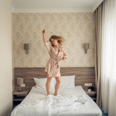 beautiful female droping pilows and jumping with bathrobe on bed in hotel room