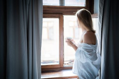 Zarif bornozlu kadın otel odasında kahve içiyor ve pencerenin yanında duruyor. Pencere yansıması