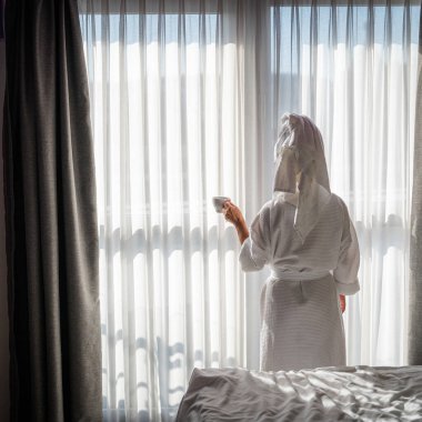 Bornozlu kadın pencerenin yanında durup pencereden dışarı bakıyor.