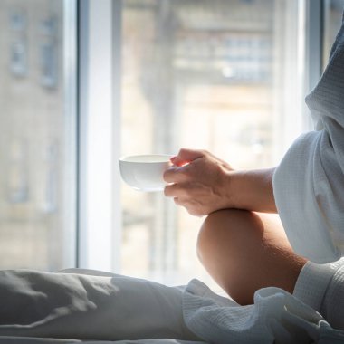 Bornozlu kadın otelde bir fincan kahveyle yatakta oturuyor.