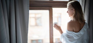 Zarif bornozlu kadın otel odasında kahve içiyor ve pencerenin yanında duruyor. Pencere yansıması