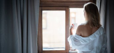 Zarif bornozlu kadın otel odasında kahve içiyor ve pencerenin yanında duruyor. Pencere yansıması